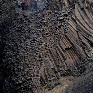 Pode incluir: Um close-up de uma formação rochosa cinza escura com uma estrutura columnar. A rocha está desgastada e tem uma textura áspera.