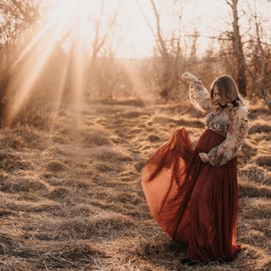 May include: A pregnant person in a long, flowing, rust-colored dress with a floral print top, standing in a field of dry grass with the sun shining through the trees in the background.