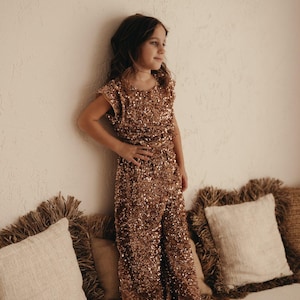 Peut inclure: Une jeune personne portant une combinaison en sequins dorés. La combinaison a des manches courtes et un col rond. La personne est debout sur une surface blanche avec des coussins derrière elle.