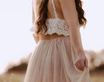May include: A woman wearing a white lace crop top and a flowing blush-colored skirt. She is wearing a white hat and has long brown hair.