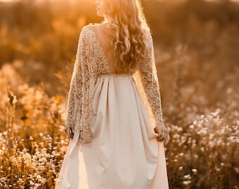 May include: A long, flowing ivory gown with a beaded bodice and sheer sleeves. The dress features a low back and a full skirt, perfect for a special occasion. The image is set in a field of wildflowers, bathed in warm sunlight.