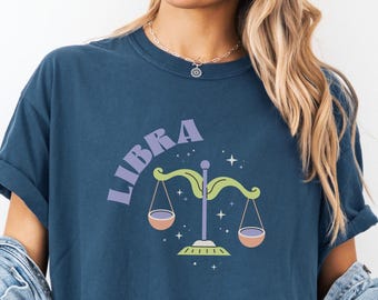 May include: A navy blue t-shirt featuring the word "LIBRA" in purple above a colorful scale design. The scales are green and the bowls are peach. The shirt is part of a zodiac-themed collection.