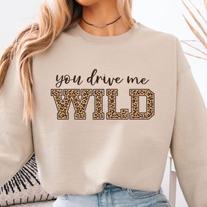 Puede incluir: Sudadera beige con el texto "you drive me WILD" en negro y estampado de leopardo. La parte superior de manga larga está hecha de un material suave. La sudadera es de estilo casual.