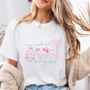 Puede incluir: Una camiseta blanca de manga corta con un estampado gráfico en rosa y turquesa. El diseño incluye un bikini, una botella de agua, gafas de sol, dos corazones y una palmera, con el texto "can I pull you for a chat?" en letra cursiva.