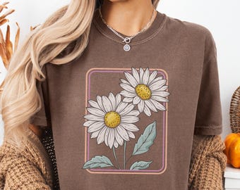 Op de afbeelding: Een bruin T-shirt met een madeliefjesafbeelding in een rechthoekige lijst. De madeliefjes hebben witte bloemblaadjes en gele centra, met groene bladeren en stelen. Het shirt heeft een relaxte pasvorm.