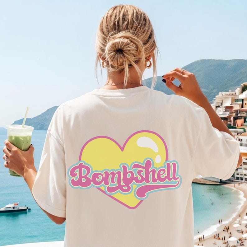 K&ouml;nnte beinhalten: Ein cremefarbenes Kurzarm-T-Shirt mit einem gro&szlig;en gelben Herz-Motiv auf der R&uuml;ckseite. Das Herz ist rosa umrandet und enth&auml;lt das Wort "Bombshell", das in einer stilisierten, kursiven rosa Schrift mit hellblauem Rand geschrieben ist.
