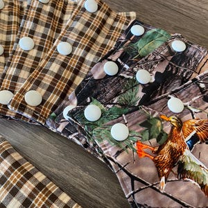 May include: A collection of fabric strips featuring white plastic snap buttons. Some strips have a brown and tan plaid pattern, while others display a camouflage print with green leaves and a flying mallard duck with orange feet.
