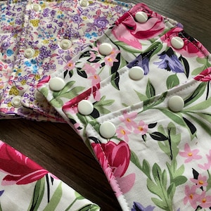 May include: A collection of reusable cloth pads featuring white fabric with floral patterns in pink, purple, and green. Each pad is rectangular with rounded corners and includes white plastic snap fasteners along the edges for secure closure.