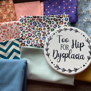 May include: A collection of colorful fabrics with various patterns, including floral, chevron, and star designs. A white, round sign with the text "Too Hip For Dysplasia" is centered in the image. The fabrics are in shades of pink, blue, and yellow.