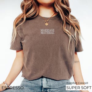 Puede incluir: Una camiseta marr&oacute;n con el texto "Marriage Material" en letras blancas. La camiseta est&aacute; hecha de una tela suave y c&oacute;moda y tiene un corte holgado.