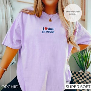 May include: A light purple t-shirt with the embroidered phrase "i ❤️ due process" in red and blue. The shirt has short sleeves and a relaxed fit. The text "EMBROIDERED DESIGN" is in a white circle. The word "ORCHID" is at the bottom left.