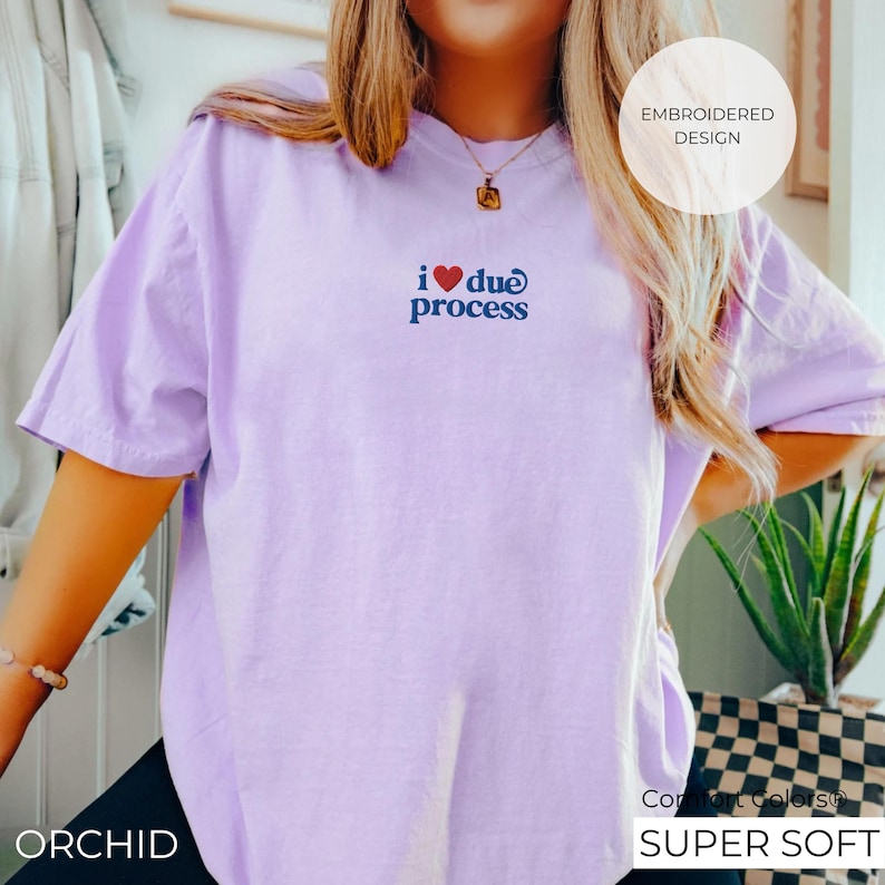 May include: A light purple t-shirt with the embroidered phrase "i ❤️ due process" in red and blue. The shirt has short sleeves and a relaxed fit. The text "EMBROIDERED DESIGN" is in a white circle. The word "ORCHID" is at the bottom left.