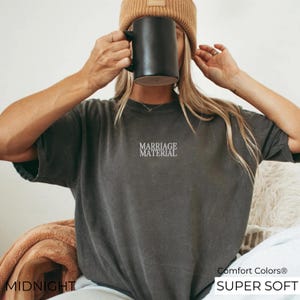 Puede incluir: Una camiseta gris oscuro con el texto "Marriage Material" bordado en la parte delantera. La camiseta est&aacute; hecha de una tela suave y c&oacute;moda y tiene un corte holgado. Comfort Colors&reg; Super Soft.