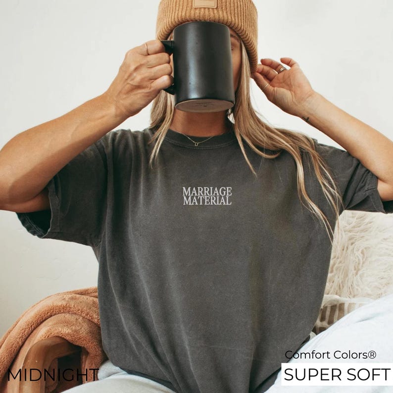 Puede incluir: Una camiseta gris oscuro con el texto "Marriage Material" bordado en la parte delantera. La camiseta est&aacute; hecha de una tela suave y c&oacute;moda y tiene un corte holgado. Comfort Colors&reg; Super Soft.