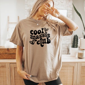 May include: A tan t-shirt with the text "cool massis club" in black with a heart design.