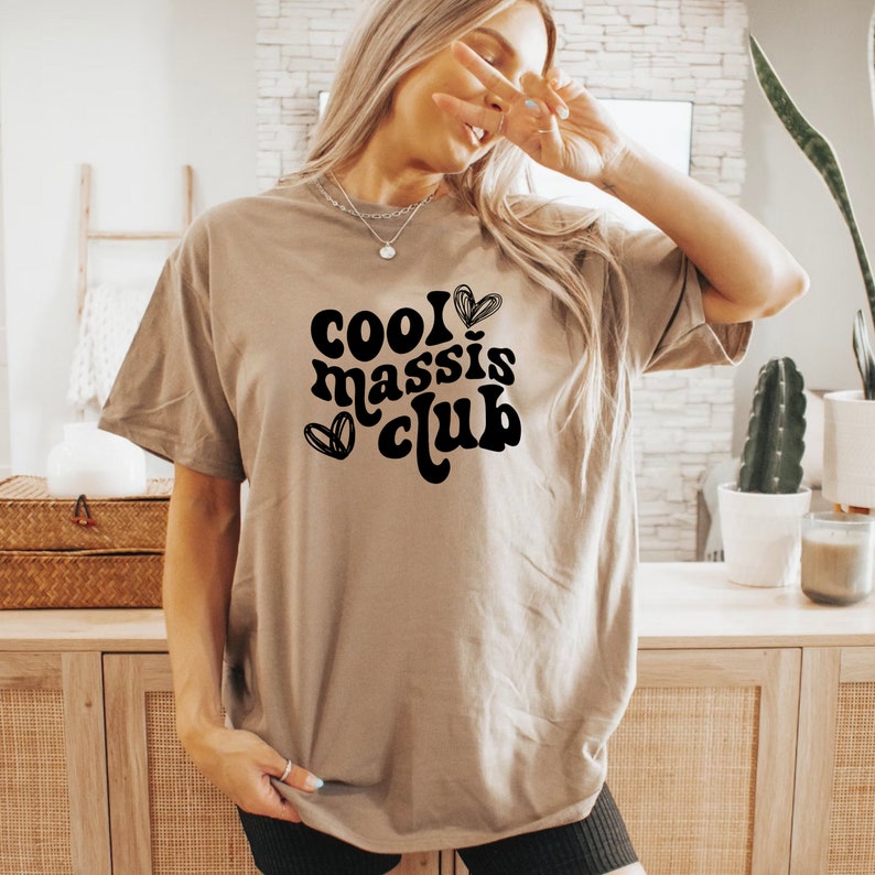 May include: A tan t-shirt with the text "cool massis club" in black with a heart design.