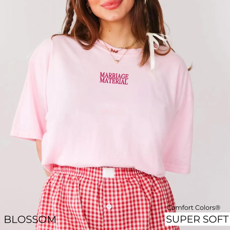 Puede incluir: Camiseta rosa de manga corta con el texto "Marriage Material" en letras rosas. La camiseta se combina con una falda de cuadros rojos y blancos.