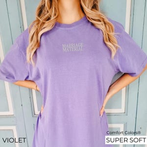 Puede incluir: Una camiseta de manga corta, s&uacute;per suave, de color morado con el texto "Marriage Material" impreso en blanco en la parte delantera.