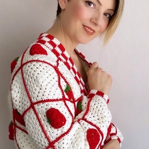 Puede incluir: Un cárdigan de crochet blanco con detalles rojos y verdes con un patrón de fresas.