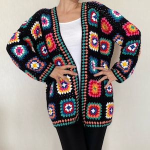 May include: A black, open-front crochet cardigan with colorful square patterns. The squares feature a variety of colors including pink, yellow, blue, and green. The cardigan has long sleeves and a decorative border.