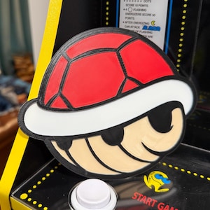 Puede incluir: Una concha roja y negra de Koopa Troopa, un personaje de la serie de videojuegos Super Mario. La concha tiene un borde blanco y está colocada sobre una superficie negra. La imagen también muestra parte de una máquina recreativa con las palabras "START GAME".