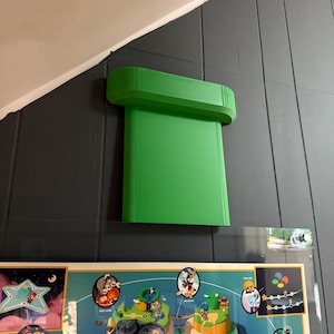 Pode incluir: Um cano verde brilhante, impresso em 3D, que lembra um videogame, está montado em uma parede cinza escura. O design do cano é um elemento reconhecível de uma popular série de videogames. Abaixo, um mapa colorido.