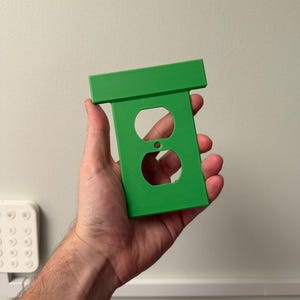 May include: A bright green electrical outlet cover with two circular openings and a rectangular top. The cover is held in a hand against a plain white wall. The cover is designed to fit over a standard electrical outlet.