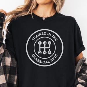 May include: A black short-sleeved t-shirt featuring a white circular graphic of a manual transmission gear shift pattern with the text "TRAINED IN THE CLASSICAL ARTS" surrounding it, worn under a black and tan plaid long-sleeved button-down shirt.