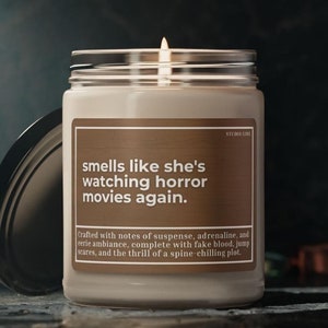 May include: A candle with a brown label that reads "smells like she's watching horror movies again." The label also includes the text "Crafted with notes of suspense, adrenaline, and eerie ambiance, complete with fake blood, jump scares, and the thrill of a spine-chilling plot."