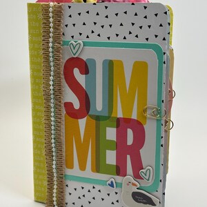 May include: A handmade summer-themed journal with the word "SUMMER" in colorful, layered letters. The journal features a white background with black triangles, a seagull sticker, and decorative ribbons. A burlap and pearl trim adorns the spine.