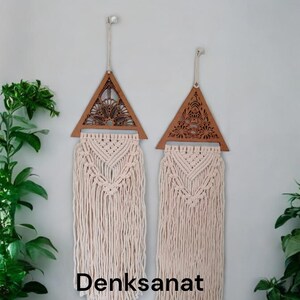 May include: Two macrame wall hangings with wooden triangle tops featuring intricate cut-out designs. The macrame is off-white, with long, flowing fringe. The text "Denksanat" is visible at the bottom. These decorative pieces add a bohemian touch to any space.