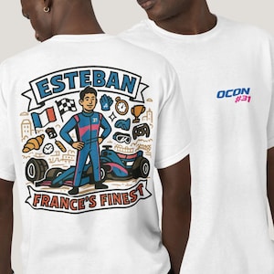 Puede incluir: Camiseta blanca con una ilustración de dibujos animados de un piloto de carreras con un traje de carreras azul y rosa, con el texto "ESTEBAN" arriba y "FRANCE'S FINEST" abajo. La camiseta también tiene el texto "OCON #31" en la parte delantera.