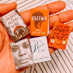 May include: A collection of five paperback books with different titles and authors. The books are all in a similar style, with colourful covers and black text. The titles include "Children of Blood and Bone", "Queenie", "How We Fight for Our Lives", "Finding Me", and "The Bluest Eye".