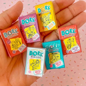Puede incluir: Cinco libros de bolsillo en miniatura con portadas coloridas que muestran el título "Dork Diaries" e ilustraciones de chicas. Cada libro tiene una pegatina con el texto "Tales from a Not-So..." y una frase diferente.