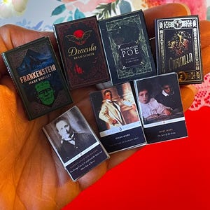 May include: A collection of miniature classic literature books, including "Frankenstein," "Dracula," and works by Edgar Allan Poe and Oscar Wilde. The books are held in a hand, with titles and author names visible on the covers. The background is a mix of floral patterns and red fabric.