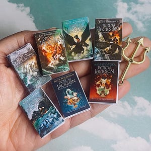 May include: A collection of seven miniature paperback books featuring the Percy Jackson series by Rick Riordan. The books are arranged in a hand, with the titles visible. The books are in various shades of blue, green, and orange, with illustrations of mythical creatures and characters.