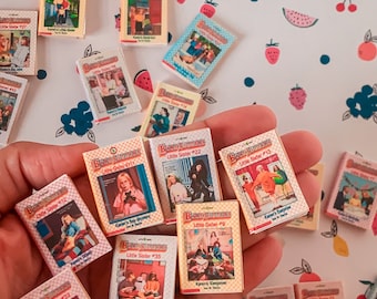 May include: A collection of miniature paperback books with colorful covers featuring the title "Baby-Sitters Little Sister" and different numbers. The books are arranged in a hand, with the covers facing the viewer. The books are all in the same style, with a white background and colorful illustrations.