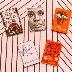 May include: Five paperback books with different covers. The books are on a white and brown striped surface. The book titles are "Children of Blood and Bone", "Finding Me", "Queenie", "The Bluest Eye", and "How We Fight for Our Lives".