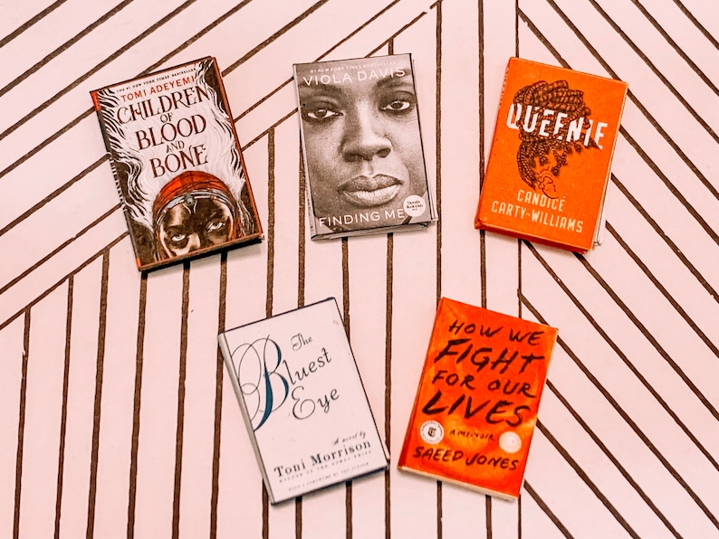 May include: Five paperback books with different covers. The books are on a white and brown striped surface. The book titles are "Children of Blood and Bone", "Finding Me", "Queenie", "The Bluest Eye", and "How We Fight for Our Lives".