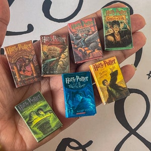 May include: A collection of miniature Harry Potter book replicas. The books feature colorful covers with titles like "Harry Potter and the Sorcerer's Stone" and "Harry Potter and the Goblet of Fire." The books are held in a hand against a white background.