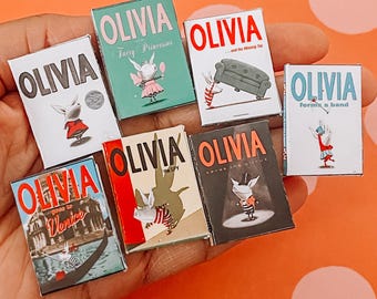 Könnte beinhalten: Eine Sammlung von Miniatur-Buchcovern von "Olivia", mit Titeln wie "Fairy Princesses" und "Forms a Band". Die Cover zeigen Illustrationen der Figur Olivia in verschiedenen Szenarien, mit einer Mischung aus Farben und Designs.