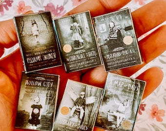 Könnte beinhalten: Eine Sammlung von sechs Miniatur-Taschenbüchern mit schwarz-weißen Covern, die Illustrationen von Kindern zeigen. Die Buchtitel lauten "Miss Peregrines Heim für Besondere Kinder", "Hollow City", "Die Konferenz der Vögel", "Bibliothek der Seelen", "Eine Karte der Tage" und "Hollows", alle von Ransom Riggs.