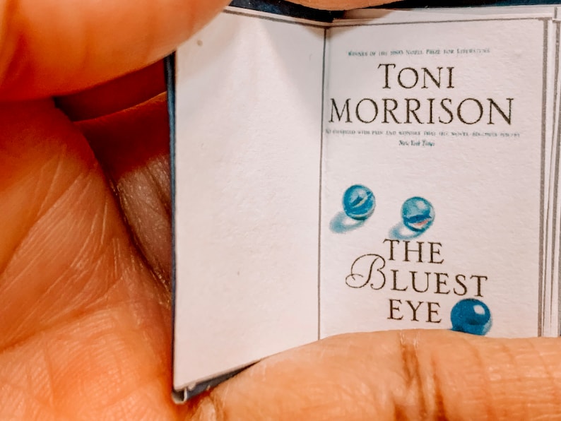 May include: A paperback book titled "The Bluest Eye" by Toni Morrison. The book cover features two blue marbles and a third blue marble on the bottom right corner.