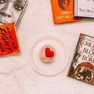 May include: A white plate with a pink cupcake topped with a red heart. The cupcake is surrounded by four books with titles including "How We Fight for Our Lives", "Children of Blood and Bone", "Candice Carty-Willi", and "A novel by Toni Morrison".