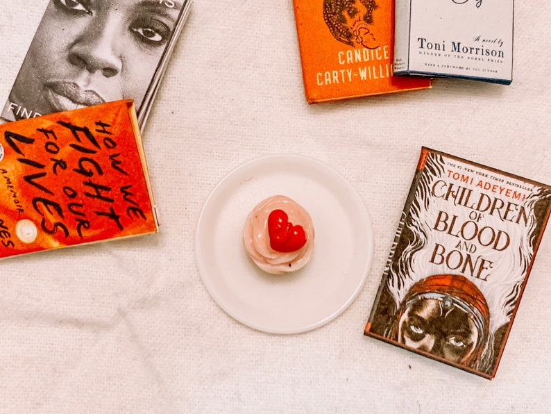 May include: A white plate with a pink cupcake topped with a red heart. The cupcake is surrounded by four books with titles including "How We Fight for Our Lives", "Children of Blood and Bone", "Candice Carty-Willi", and "A novel by Toni Morrison".