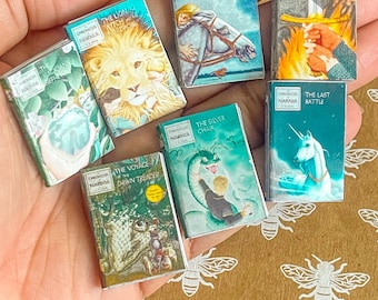 Könnte beinhalten: Eine Sammlung von sieben Miniatur-Hardcover-Büchern mit bunten Covern, die Illustrationen aus der Reihe Die Chroniken von Narnia zeigen. Die Bücher sind in einer Hand angeordnet, wobei die Titel sichtbar sind: Der Löwe, die Hexe und der Kleiderschrank, Das Pferd und sein Junge, Prinz Kaspian, Der Silberne Sessel, Die Reise auf der Morgenröte und Die letzte Schlacht.