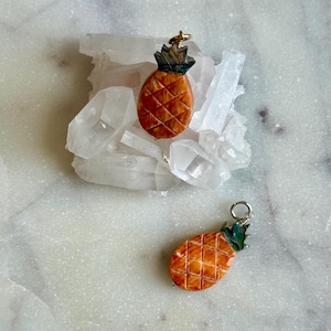 May include: Two orange pineapple pendants with green and brown tops. The pendants are set against a background of clear quartz crystals. The pineapples have a textured surface and are ready to be worn as jewelry.