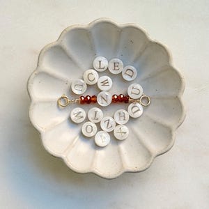 May include: A handmade bracelet with gold-colored wire and small, round, white letter beads spelling out the word "WONDER". The bracelet features a central section of small, red-brown beads. The bracelet is displayed in a white, scalloped dish.
