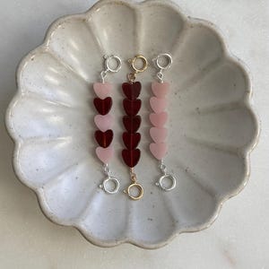 May include: Three beaded bracelets with heart-shaped beads in shades of pink and red. Each bracelet has a different clasp color: silver, gold, and silver. The bracelets are displayed on a decorative, scalloped-edge ceramic dish.