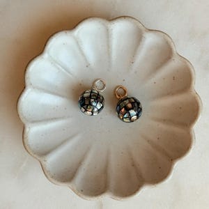 May include: Two round, mosaic-style charms with iridescent black, silver, and gold tones. One charm has a silver-colored loop, the other a gold-colored loop. They rest on a light beige, scalloped-edge ceramic dish, set against a marble background.